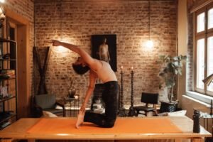 Side view full body of slim barefoot woman in sportswear doing Ushtrasana or Camel asana on yoga mat on wooden platform with arm raised in light room